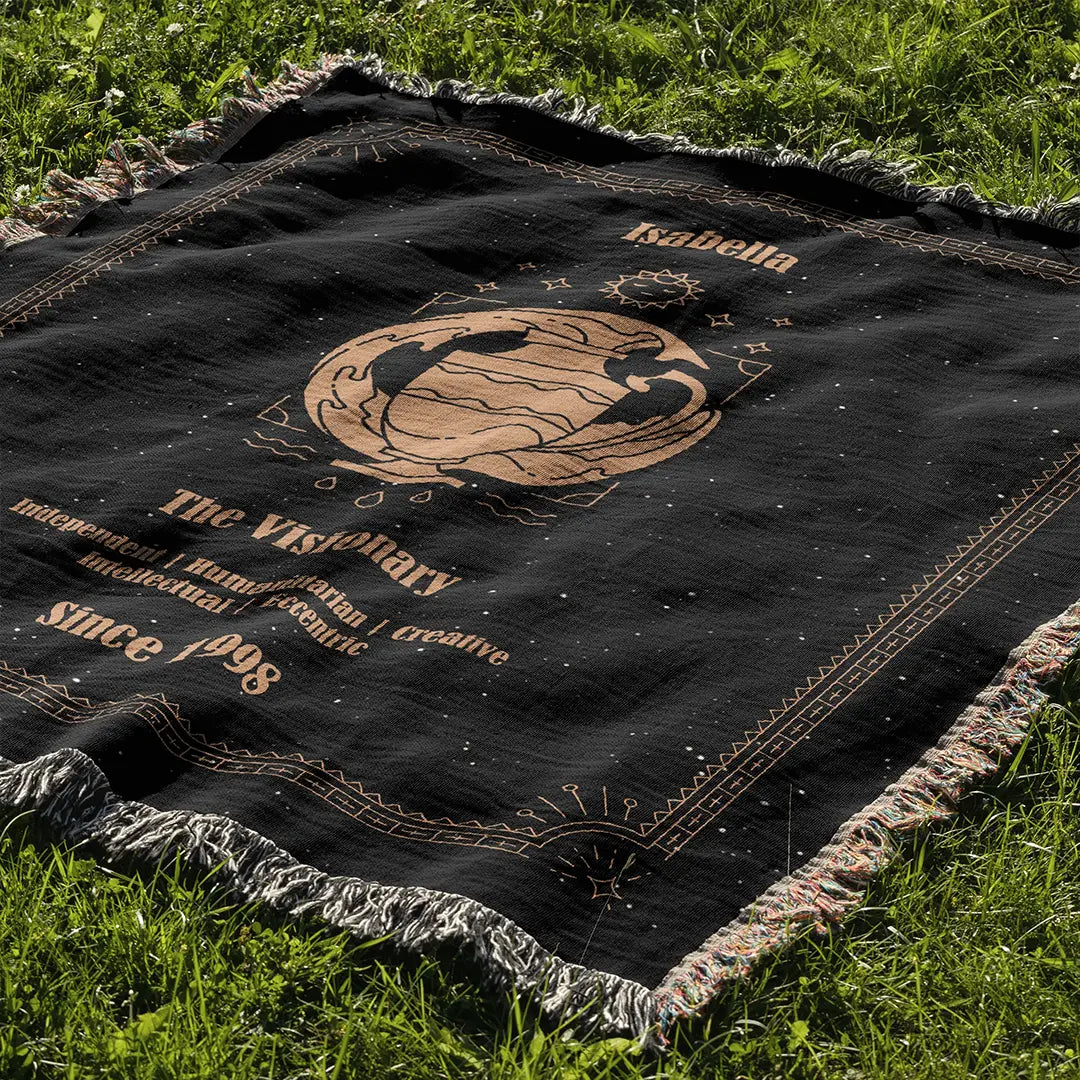 Black Aquarius zodiac-style custom woven cotton blanket laid out on a sunlit green lawn, displaying personalized name and traits.