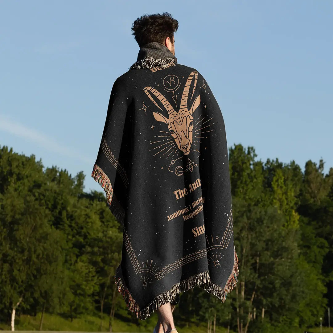 Man wrapped in a Capricorn-themed custom woven cotton blanket with horned goat artwork, standing in a grassy field under a clear sky.