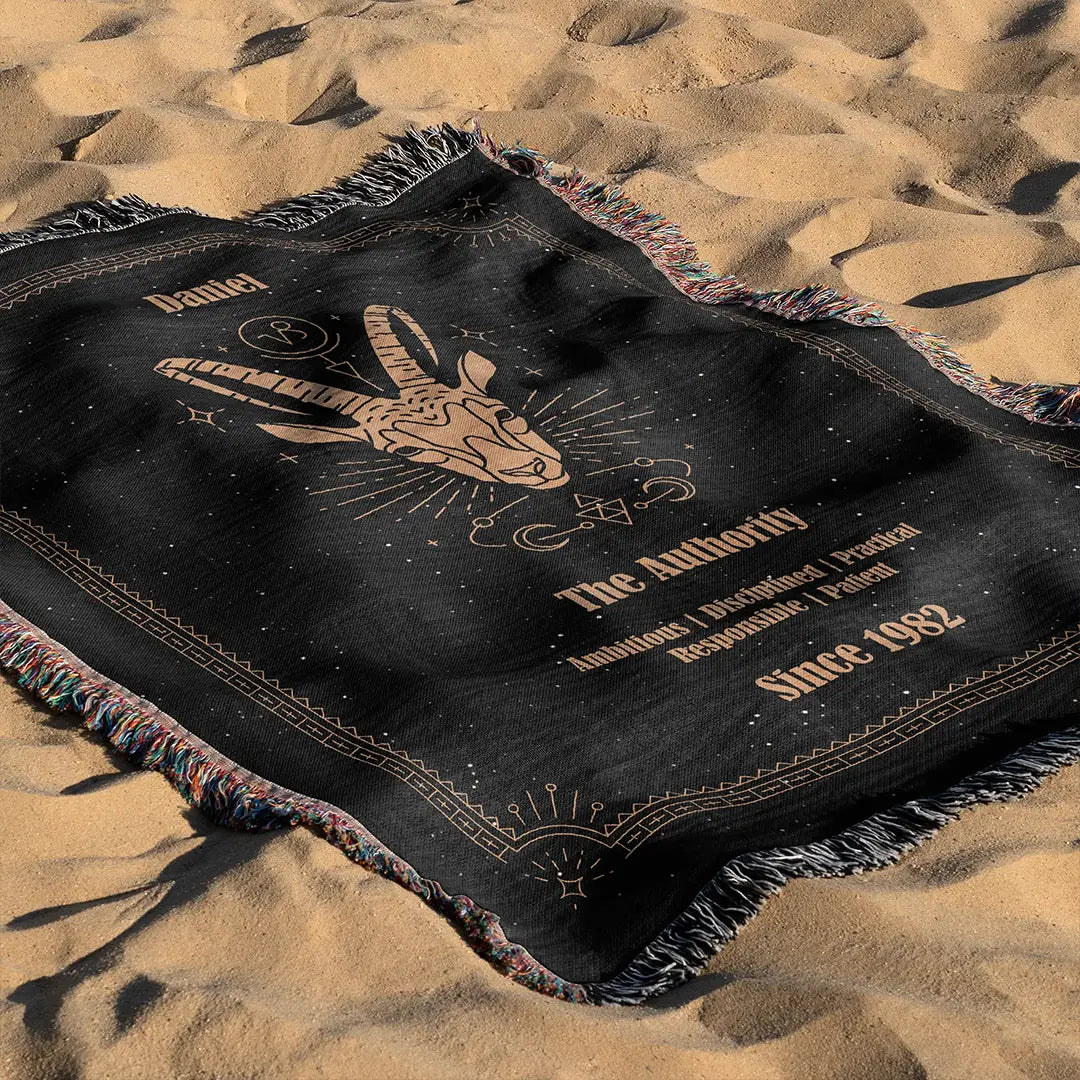 Capricorn custom woven cotton blanket laid out on golden beach sand, displaying the goat head symbol and astrology motifs under natural light.