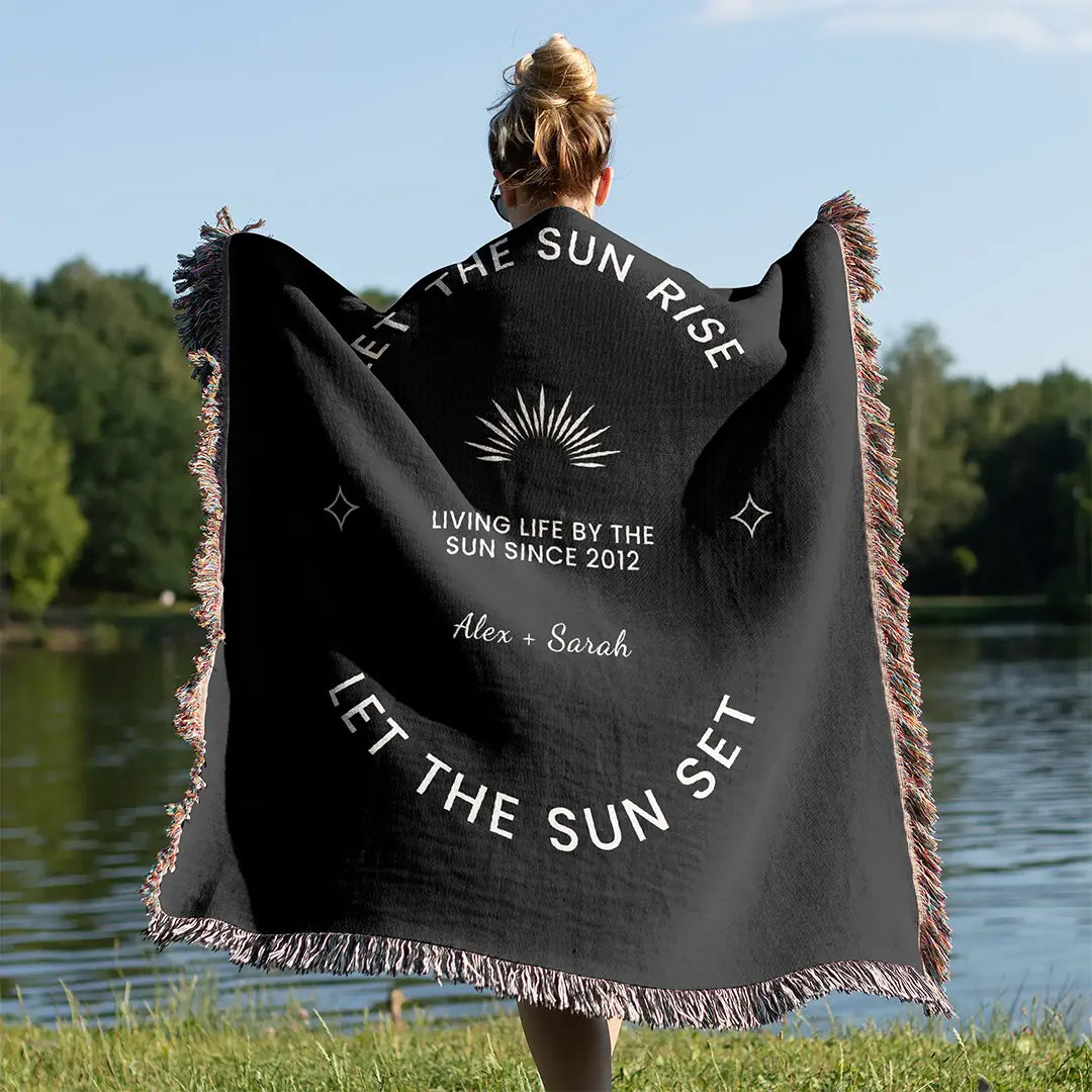 A woman stands wrapped in a black custom woven cotton name blanket for couples with a radiant sun and personalization text options.