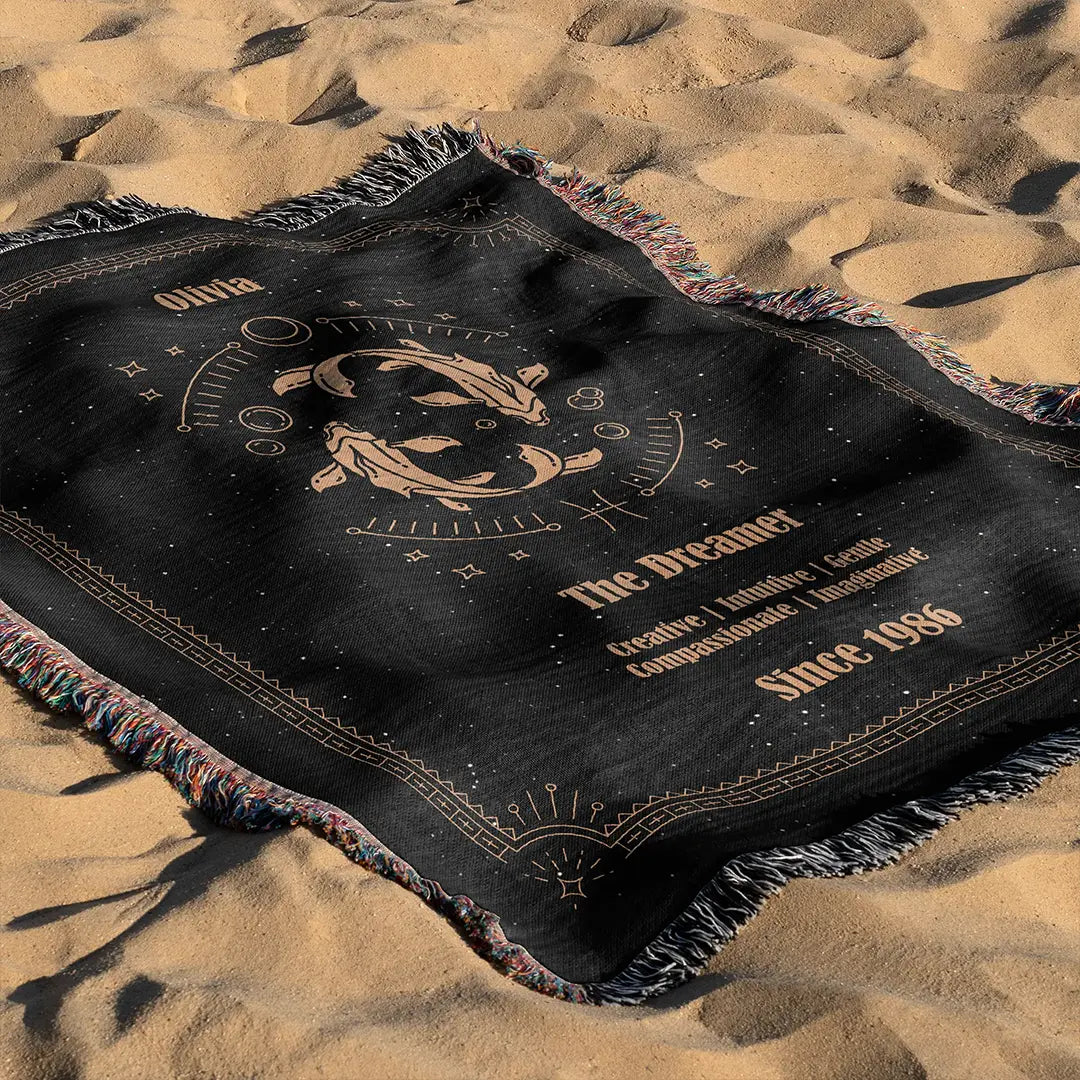 A Pisces custom woven cotton blanket laid out on soft beach sand. The celestial fish design and decorative border pop against the black fabric.