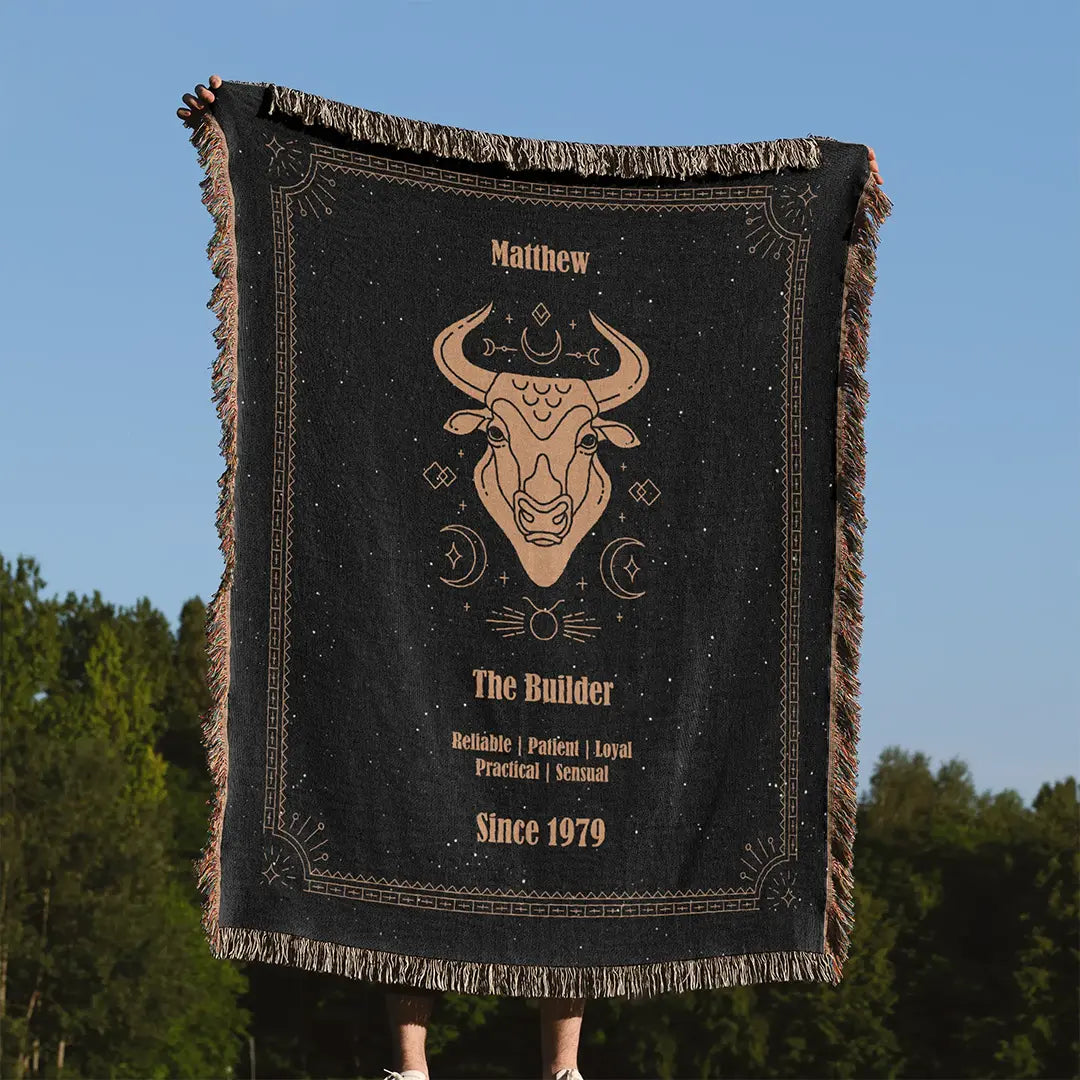 A custom woven cotton blanket with a Taurus design featuring a custom name held upright against a clear blue sky with a forest in the background.