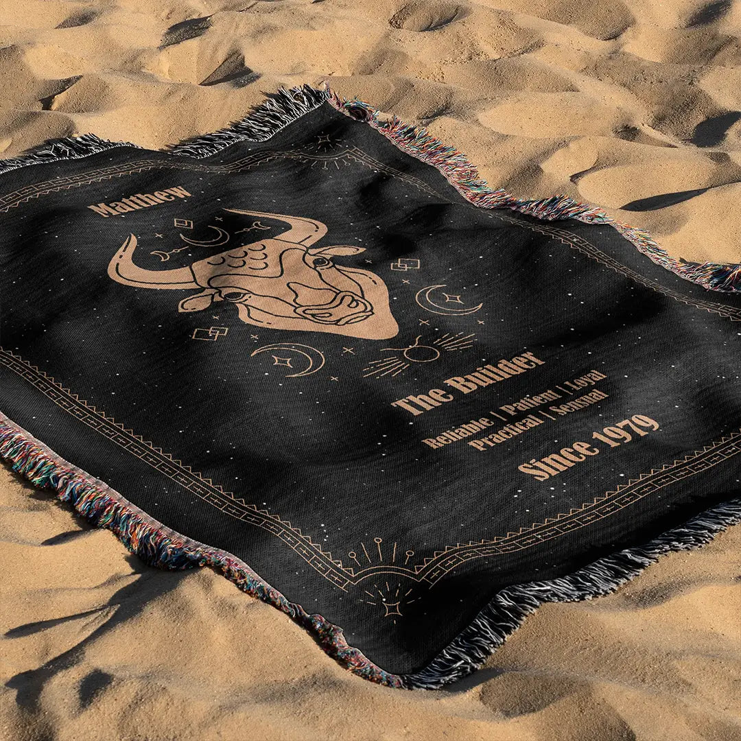 A custom woven cotton blanket displaying a bull illustration and Taurus characteristics, spread out on soft beach sand under daylight.