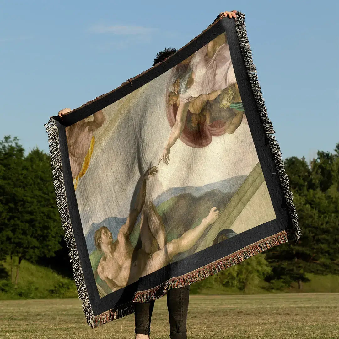 Woven cotton blanket held outdoors in a grassy field, featuring The Creation of Adam by Michelangelo, with black fringe edges and full artwork detail.