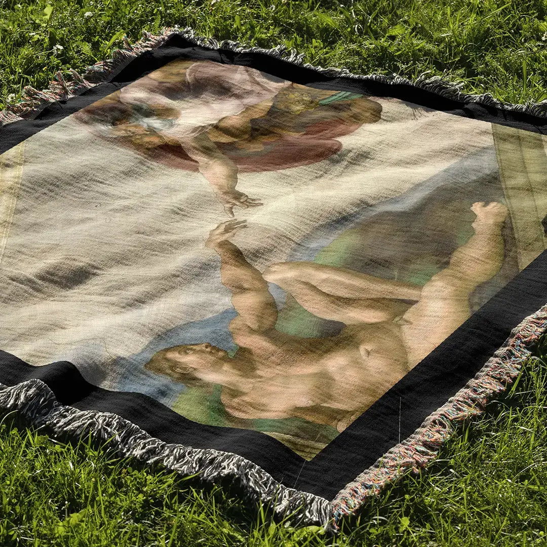 Woven cotton blanket laid out on green grass, displaying The Creation of Adam by Michelangelo, with full illustration and multicolor fringe trim.