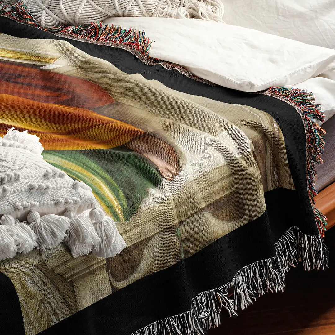 Michelangelo’s "Delphic Sibyl" woven blanket displayed on a bed with white pillows and a textured knit throw. Vibrant colors and intricate weave detail visible.