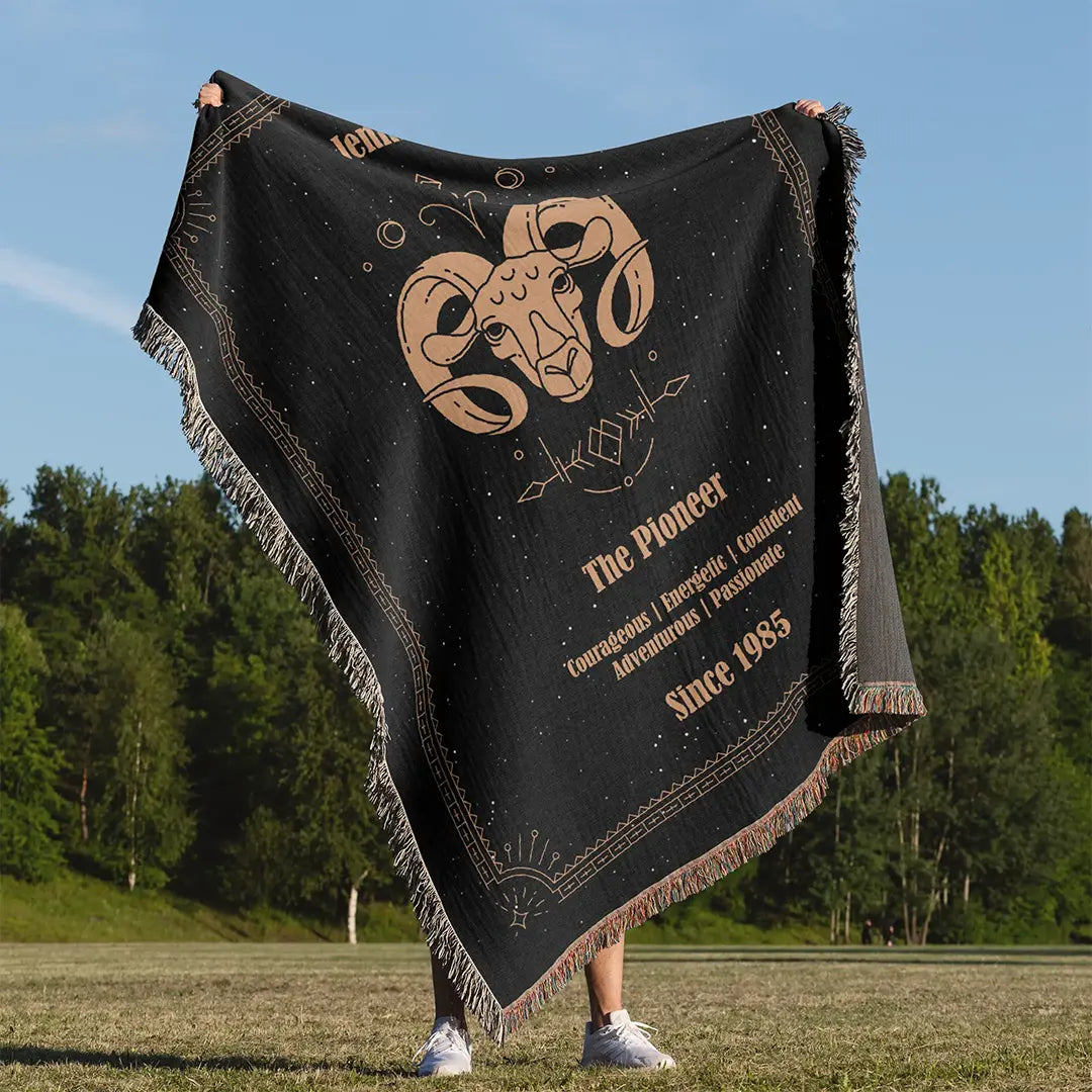 A custom woven cotton blanket featuring the Aries zodiac design, held up outdoors against a forest backdrop on a sunny day.