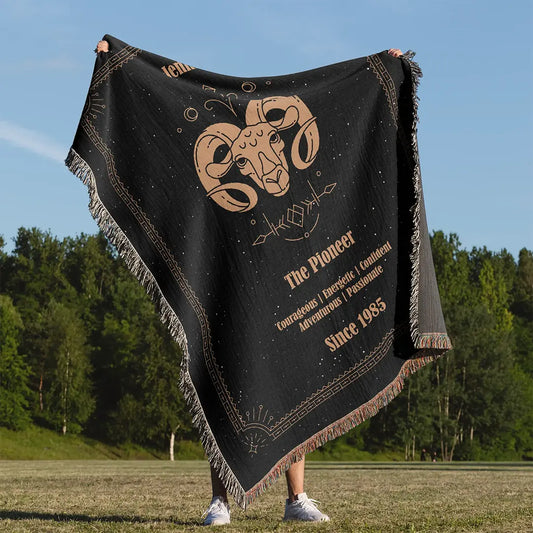 A custom woven cotton blanket featuring the Aries zodiac design, held up outdoors against a forest backdrop on a sunny day.