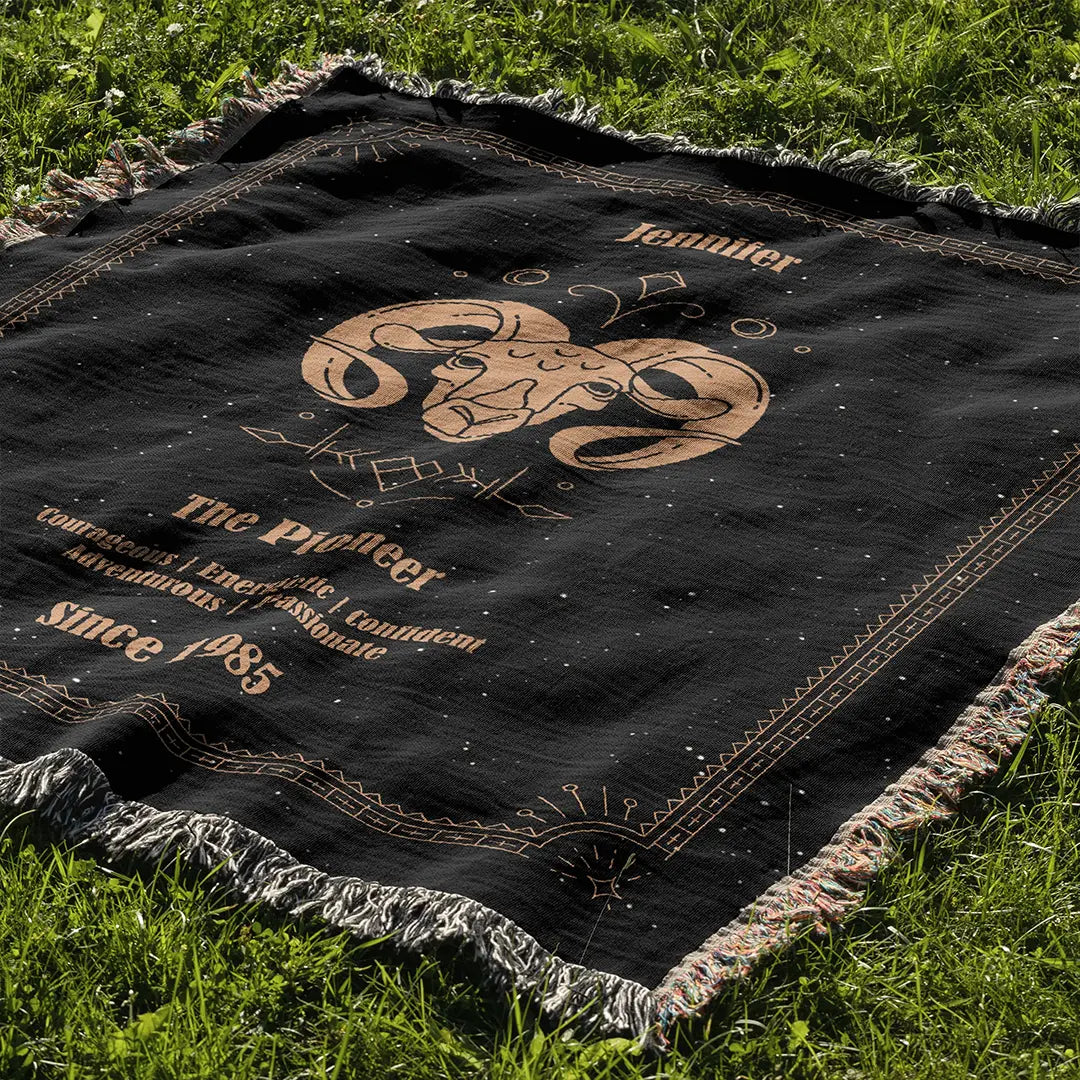 Custom woven cotton blanket laid on green grass, presenting a centered Aries motif with zodiac symbols and intricate border details.