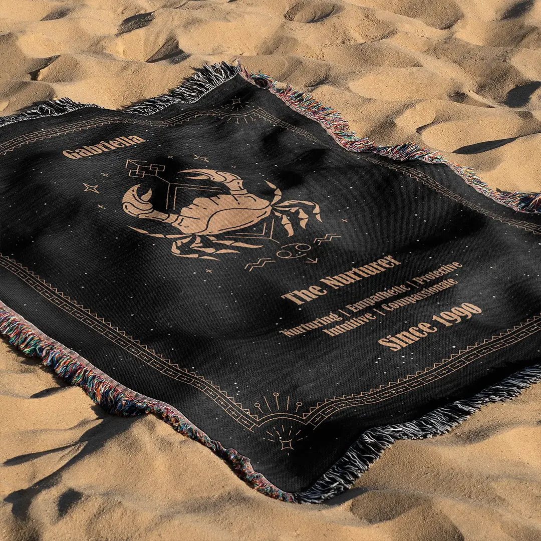 Custom woven cotton blanket showcasing a Cancer Zodiac design spread out on golden beach sand.