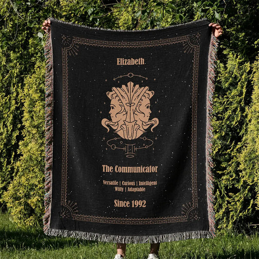 Custom woven cotton blanket featuring a Gemini zodiac design, held up outdoors against vibrant green foliage.