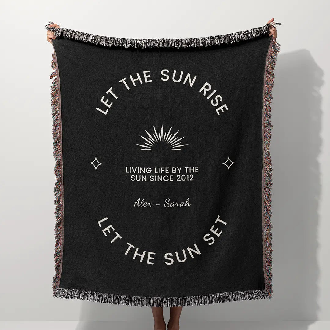 A woman stands wrapped in a black custom woven cotton name blanket for couples with a radiant sun and personalization text options.