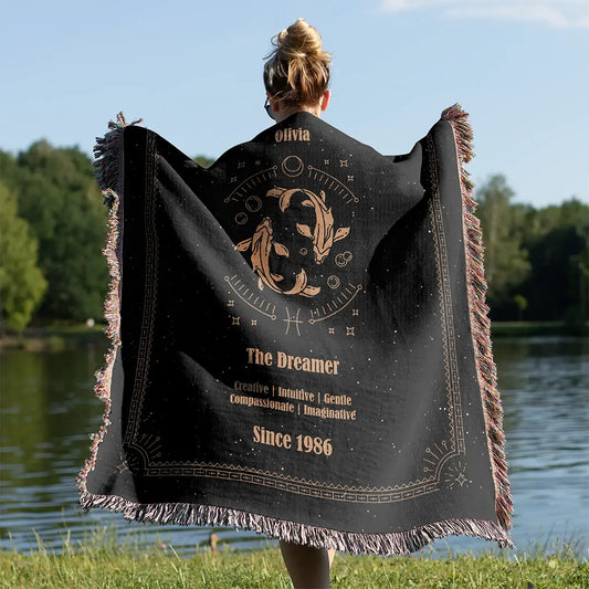 A woman wrapped in a custom woven cotton blanket featuring a Pisces zodiac sign, standing near a calm lake under clear skies.