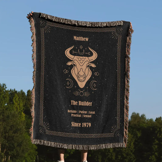 A custom woven cotton blanket with a Taurus design featuring a custom name held upright against a clear blue sky with a forest in the background.