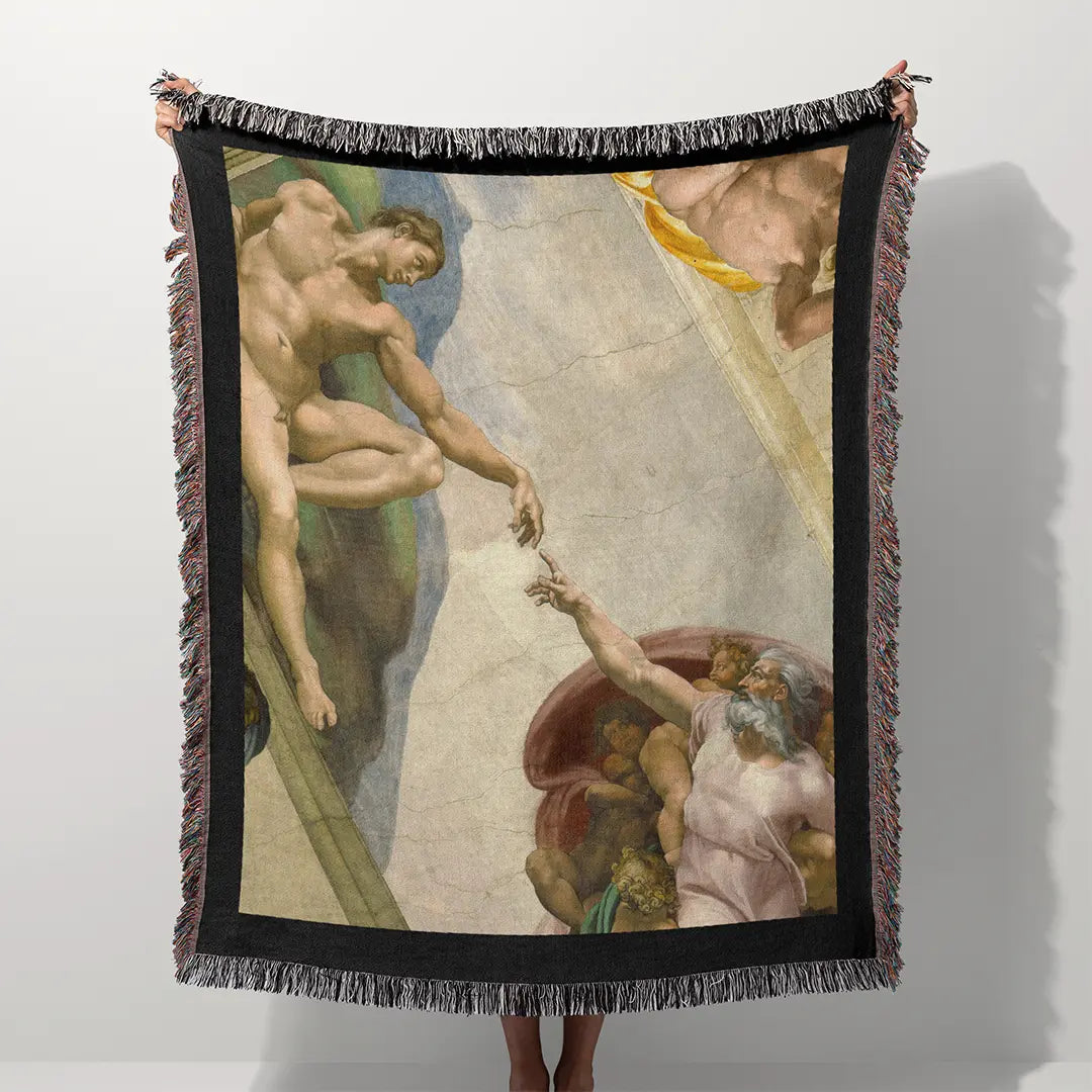 Woven cotton blanket held outdoors in a grassy field, featuring The Creation of Adam by Michelangelo, with black fringe edges and full artwork detail.