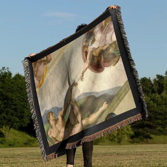 Woven cotton blanket held outdoors in a grassy field, featuring The Creation of Adam by Michelangelo, with black fringe edges and full artwork detail.
