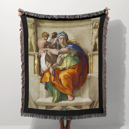 Woven blanket featuring Michelangelo's "Delphic Sibyl" design, held outdoors with trees and sky in the background. Multicolor fringe edges are visible.