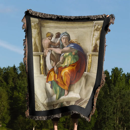 Woven blanket featuring Michelangelo's "Delphic Sibyl" design, held outdoors with trees and sky in the background. Multicolor fringe edges are visible.
