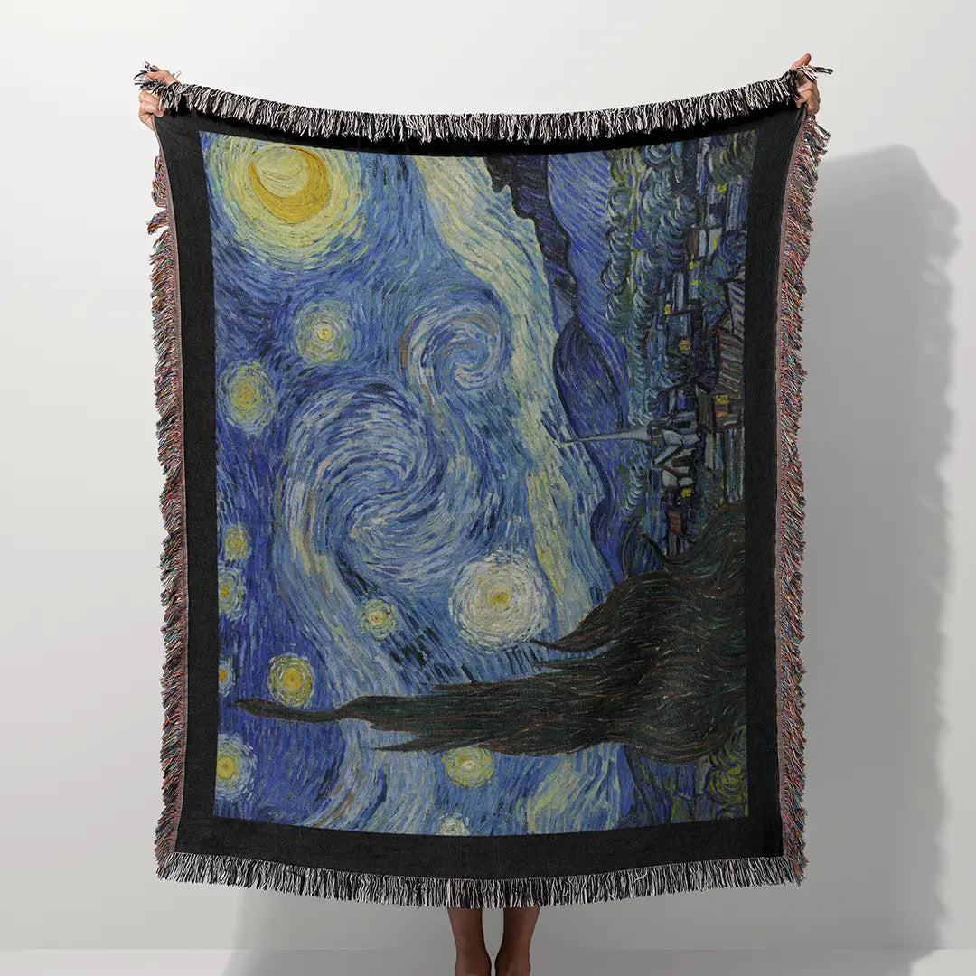 Person holding up a woven cotton blanket outdoors featuring Vincent van Gogh's "The Starry Night" design in rich blue and yellow tones, framed with black edges and colorful fringe.
