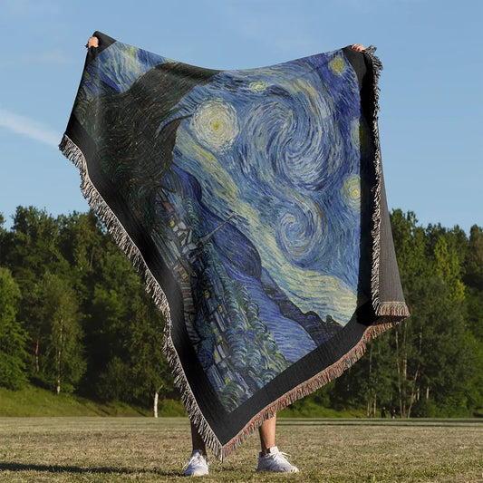 Person holding up a woven cotton blanket outdoors featuring Vincent van Gogh's "The Starry Night" design in rich blue and yellow tones, framed with black edges and colorful fringe.